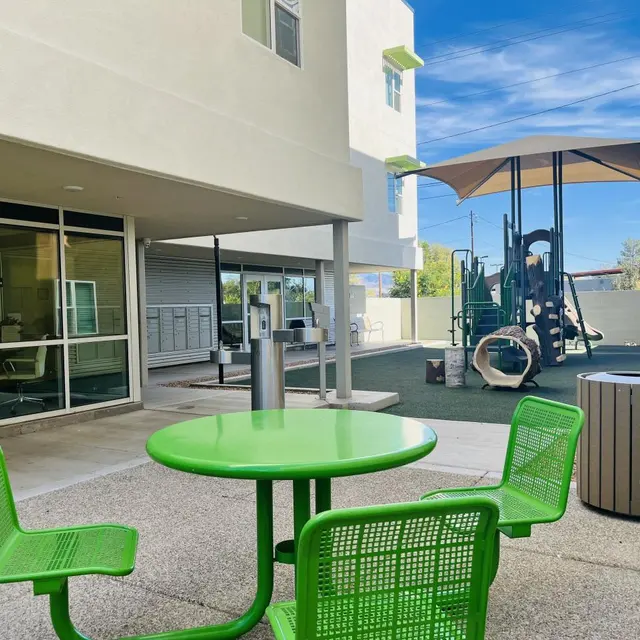 View of a playground area featuring green circular tables and chairs, a play structure in the background, and a sunny sky.