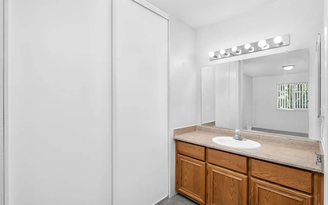A bright and clean bathroom featuring a wooden vanity with a sink and a mirror, illuminated by wall-mounted light fixtures. The walls are painted white, and there is a sliding door on one side. A window allows natural light into the space, with blinds partially open.