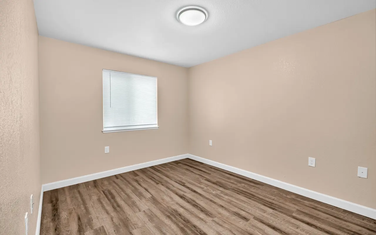 A vacant and unfurnished room with light beige walls, a window with white blinds, and a brown wooden floor.