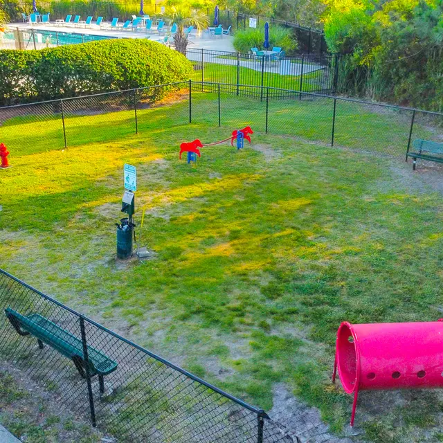 An aerial view of a fenced dog park featuring green grass, benches, a dog play equipment area, and adjacent swimming pool area in the background.