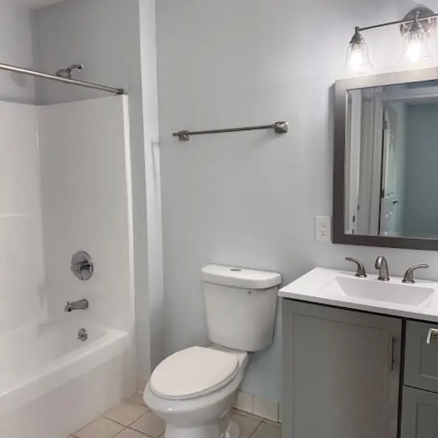 A modern bathroom featuring a white tub with a shower, a toilet, and a gray vanity with a sink. The walls are painted light blue and there is a mirror above the sink.