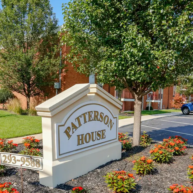 A white and beige sign displaying 'PATTERSON HOUSE' with a phone number, surrounded by well-maintained landscaping, including shrubs and flowers, and a brick building in the background.