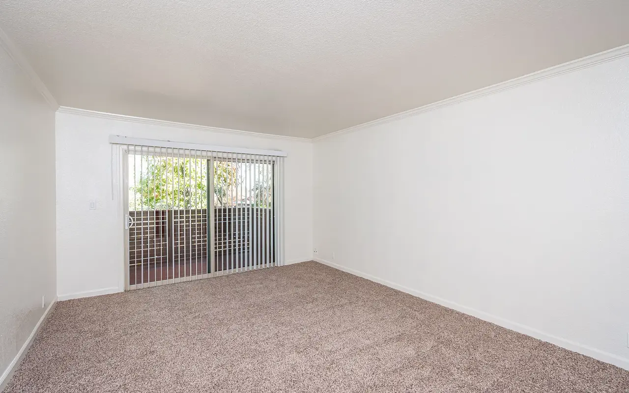 An empty living room with beige carpeting and white walls. A sliding glass door with vertical blinds leads to an outdoor balcony area.