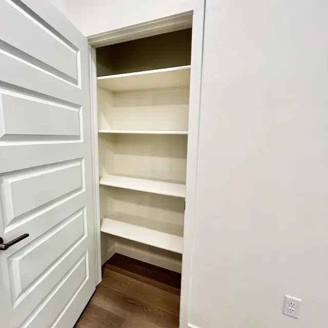 A small, modern pantry or storage closet featuring several white shelves. The door is partially open, revealing the interior.