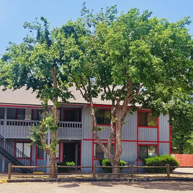 Apartment Building Surrounded by Trees A gray apartment building with red trim and stairs on the left side, surrounded by leafy trees and greenery.