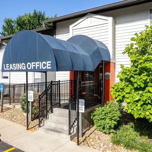 Exterior view of a leasing office with a blue canopy labeled 'LEASING OFFICE' and green shrubs in the foreground.
