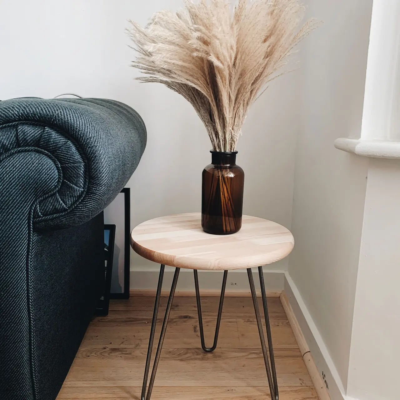 A cozy corner featuring a small wooden table with metal legs, topped with a dark amber vase containing dried pampas grass. The couch is a dark color, complementing the wooden floor and the light wall.