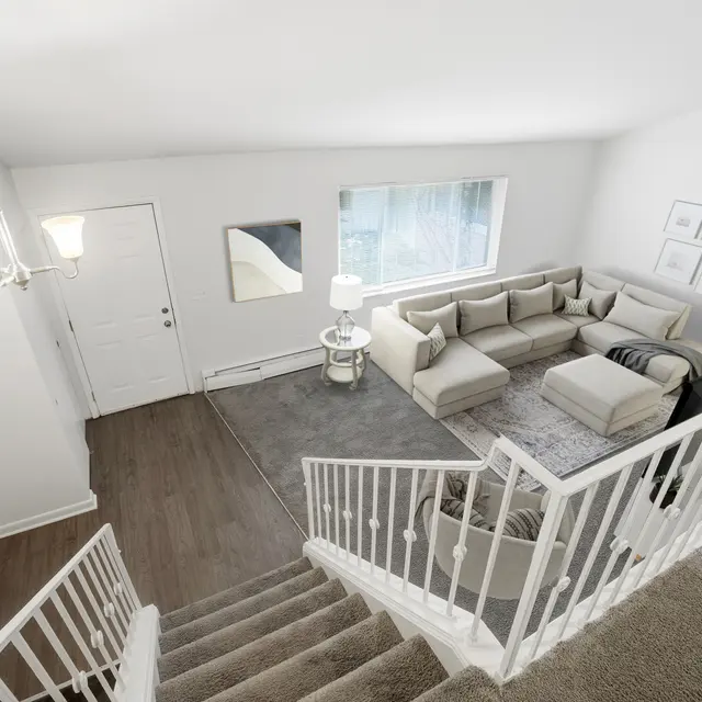 A spacious and modern living room seen from an upper angle, featuring a light-colored sectional sofa, a round coffee table, and a large window that lets in natural light. A staircase with white railings leads down into the room, and the walls are painted white with some framed art.