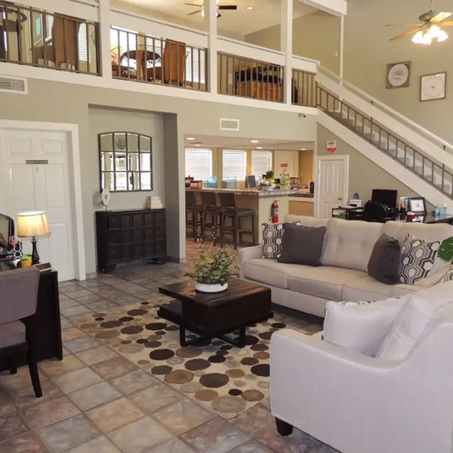 Beautiful Modern Living Room A spacious and airy living room with high ceilings, featuring a light-colored sofa, coffee table with a plant, and patterned rug. A desk with a lamp and a chair is on the left, and a staircase leads to the upper level. In the background, there is an open area with a kitchen and dining space.