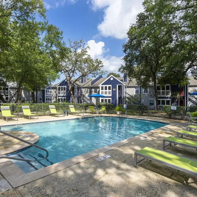 A sunny pool area with green lounge chairs, surrounded by trees and apartment buildings under a blue sky.
