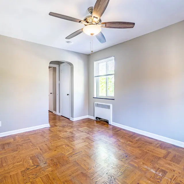 A bright, empty room featuring a ceiling fan and a parquet floor. There is a window letting in natural light and an archway leading to another space.