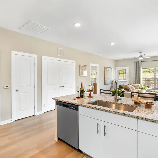 An open concept kitchen with a granite countertop and a view of a living room area that includes a couch and large windows.