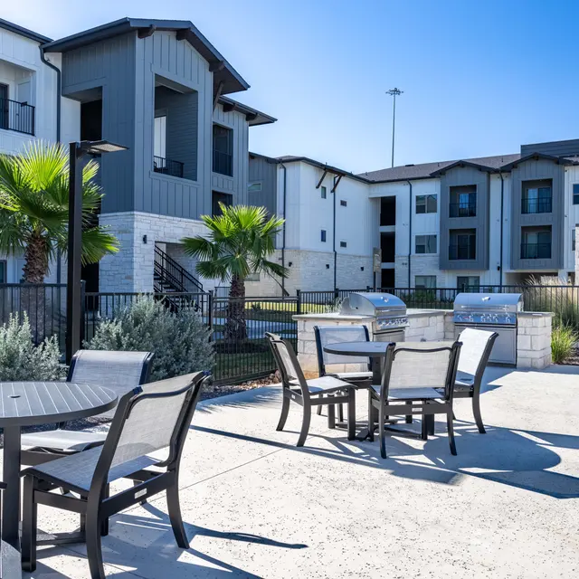 A modern apartment complex with a patio area featuring tables and chairs, surrounded by greenery and palm trees, under a clear blue sky.
