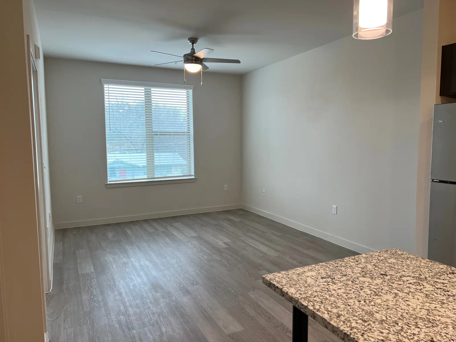 Modern Apartment Living Area A modern living area with gray laminate flooring, a ceiling fan, and a window allowing natural light. The room includes a small kitchen counter with granite surface on the right side.
