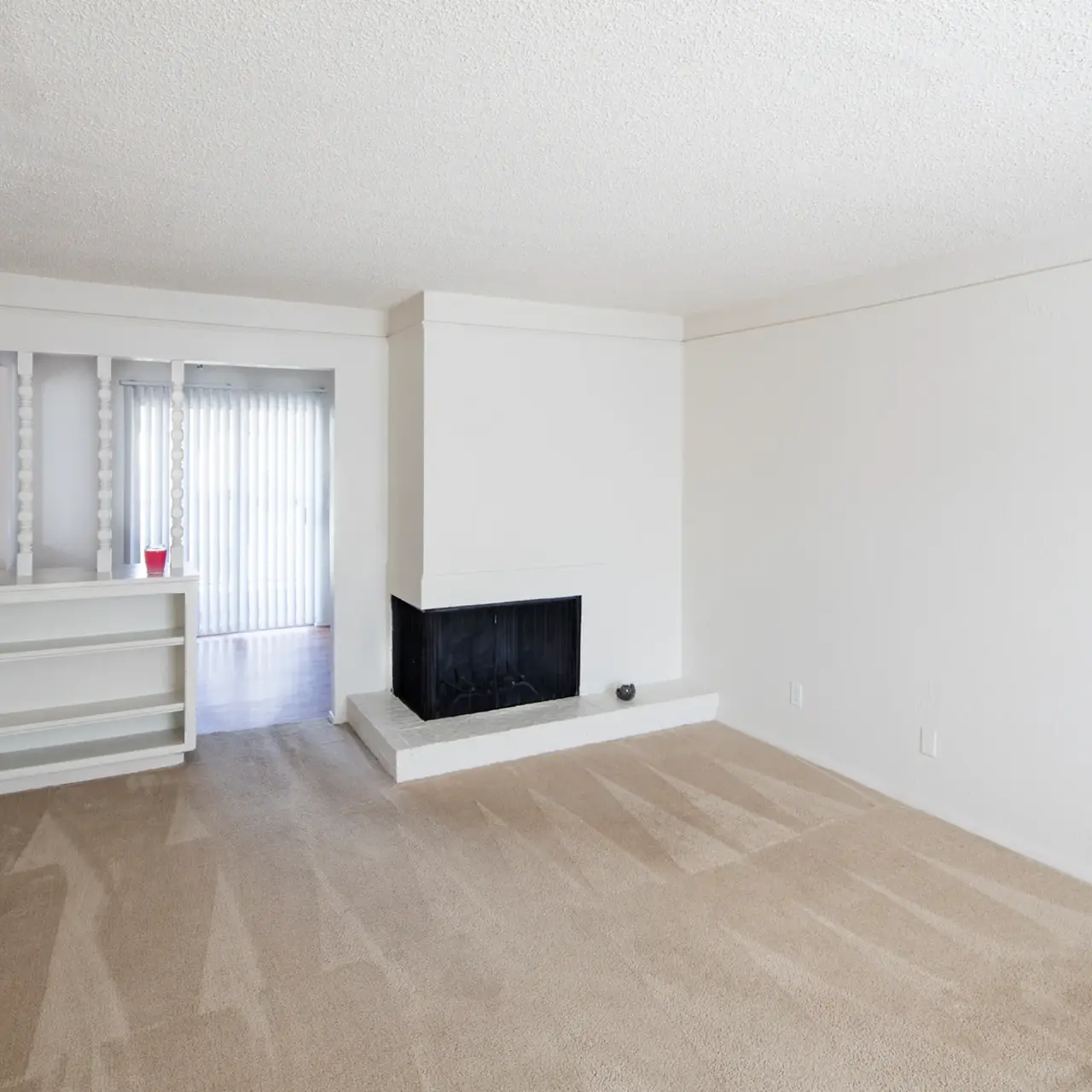 A spacious and empty living room featuring beige carpet flooring and a white-painted wall. A white fireplace is centered against one wall, and there is a small shelf in the corner. A door leads outside, and vertical blinds partially cover a window.