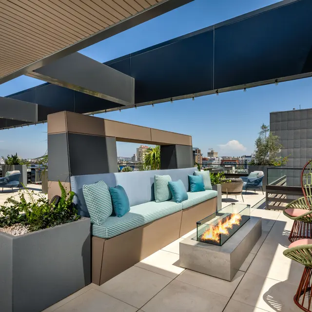Stylish rooftop lounge area featuring modern seating, a fire pit, and decorative greenery with a clear blue sky in the background.