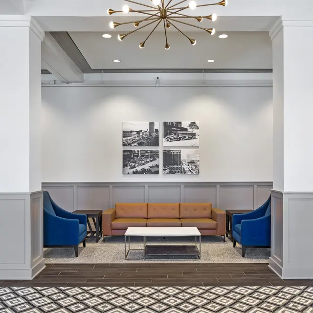 A modern lobby area featuring several blue chairs, a brown couch, and a central coffee table. The walls are adorned with framed black and white photographs. The flooring has a geometric pattern, and the space is well-lit with an artistic light fixture hanging from the ceiling.