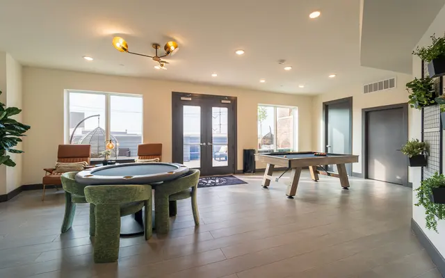 A spacious game room featuring a round poker table with green velvet chairs, two seating areas, a pool table, and large windows allowing natural light, decorated with indoor plants.