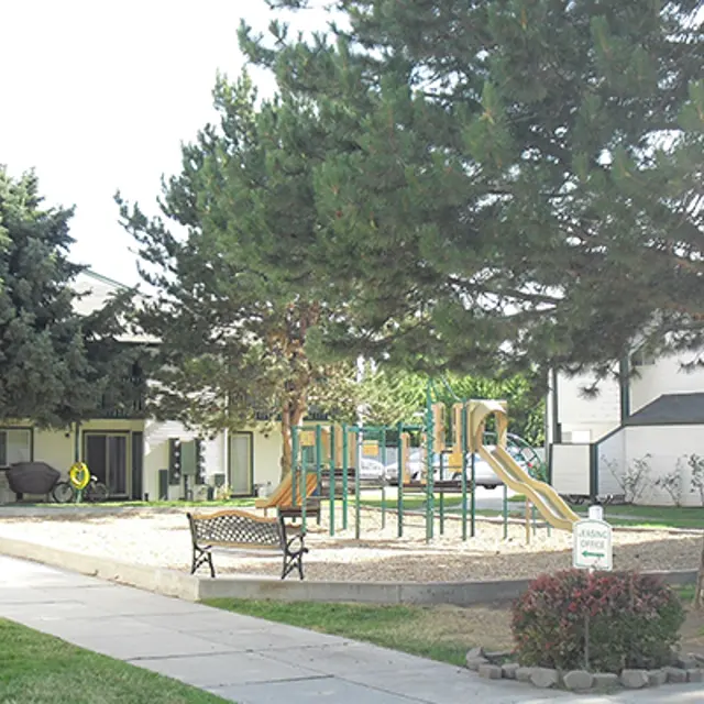 Residential Playground Area A playground area with slides and climbing structures surrounded by trees and benches, set in a residential environment with buildings in the background.