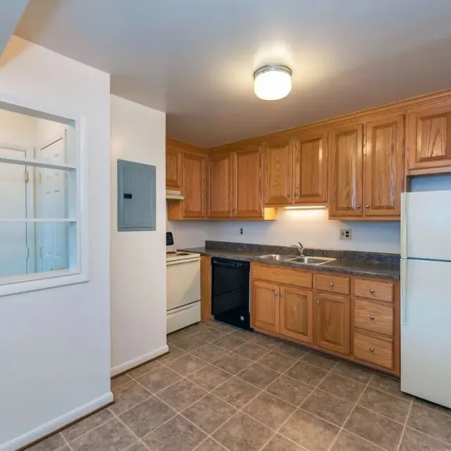 A kitchen featuring light wood cabinets, a dark countertop, and white appliances including a refrigerator and dishwasher. The floor is tiled in a neutral color, and there is an open view into another room.
