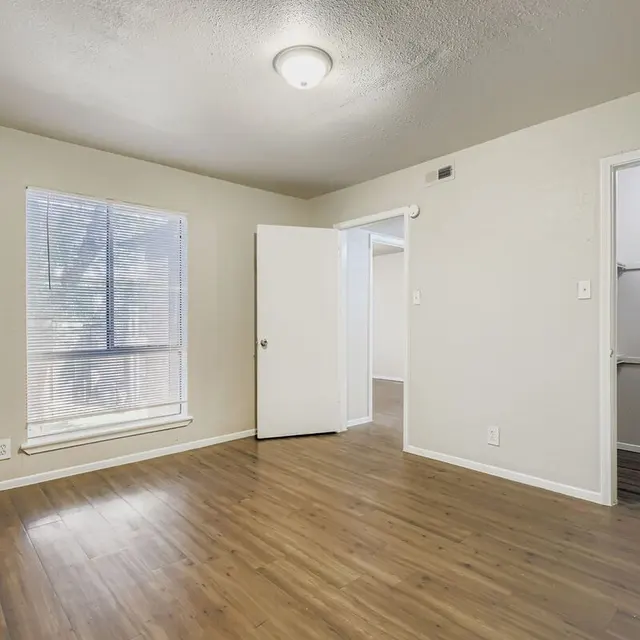 An empty room featuring light-colored walls, a window with Venetian blinds, and wooden flooring. There are two doors leading to other rooms, and a ceiling light fixture.