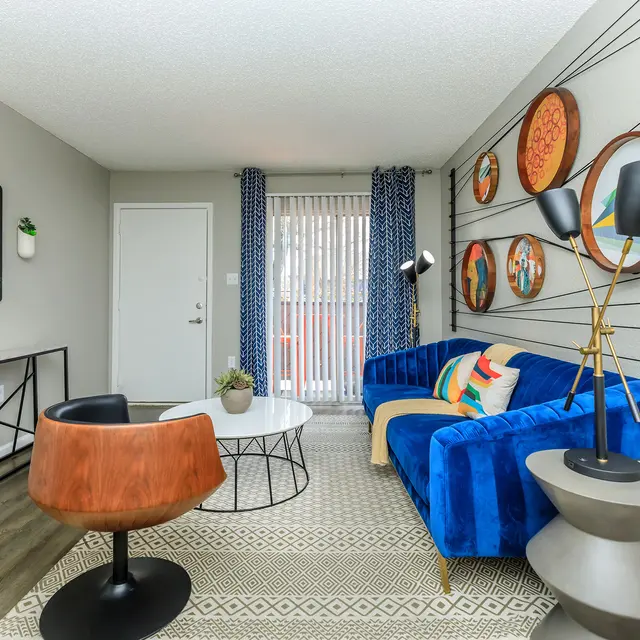 A modern living room featuring a blue sofa with colorful cushions, an orange accent chair, and a circular coffee table. There is a wall decorated with round art pieces and a patterned rug on the floor.