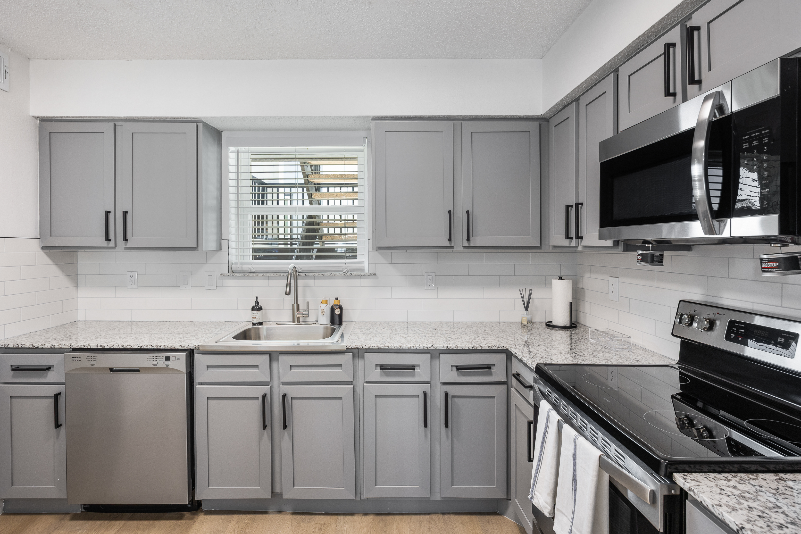 A modern kitchen featuring gray cabinets, stainless steel appliances, and a white tiled backsplash. There is a sink under a window and a granite countertop.