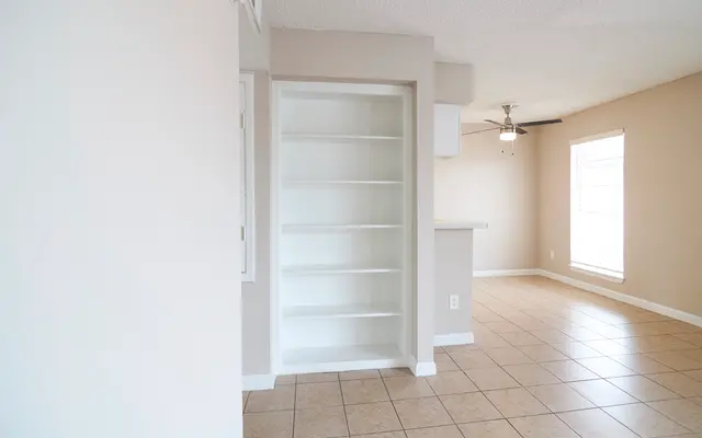 A view of a light and airy interior space featuring beige walls and a tiled floor. There is a built-in white bookshelf on the left, and a ceiling fan is visible on the right. Large windows allow natural light to fill the room.