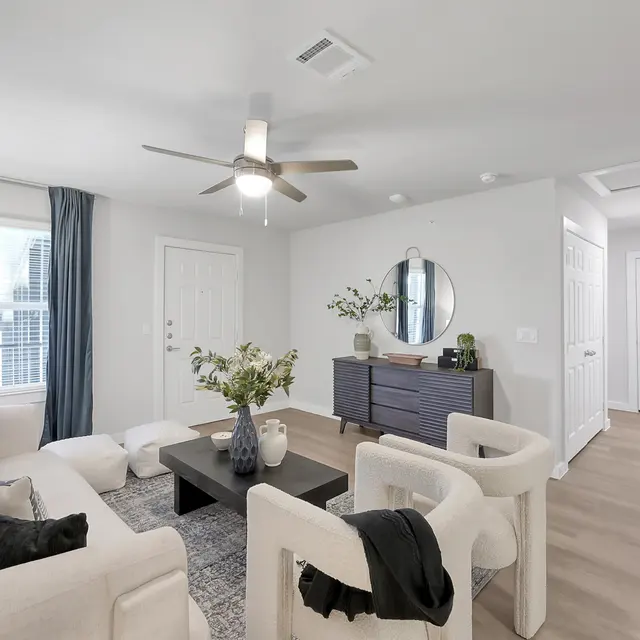 A modern living room featuring a cream-colored sofa with decorative pillows, two stylish white armchairs, and a black coffee table. The room is well-lit with natural light streaming through large windows covered with blue curtains. A round mirror hangs on the wall above a wooden sideboard, while a small green plant adds a touch of nature to the decor.