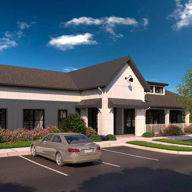A modern building with a sloped roof and light-colored exterior, surrounded by greenery and parked cars.