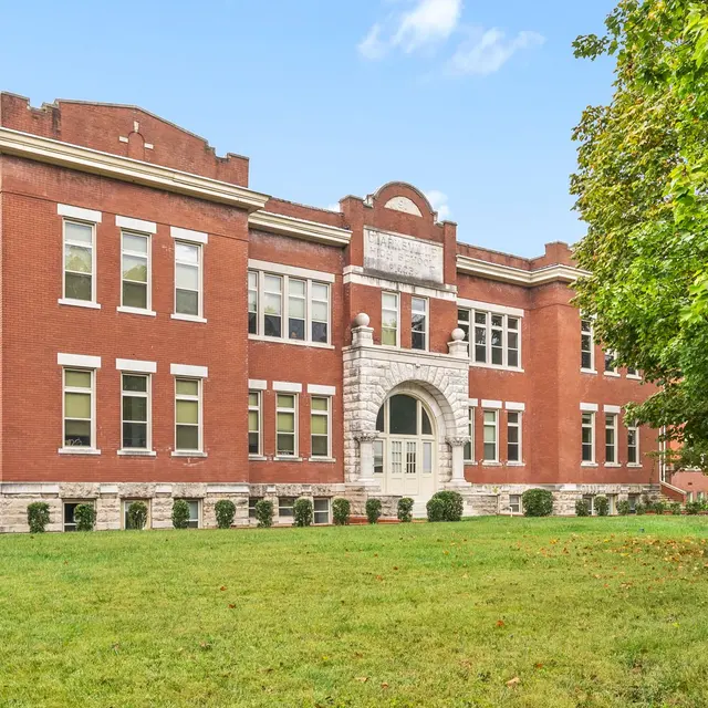 A historic red brick school building with a decorative entrance and large windows, set on a grassy lawn with a tree on the side.