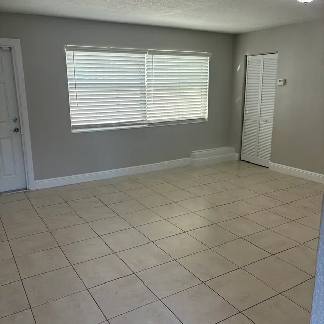 An empty living room with tiled floor, light gray walls, and a window with blinds.