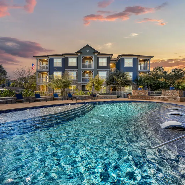 An apartment complex with a swimming pool in the foreground, reflecting the colors of a sunset. The pool is surrounded by lounge chairs and greenery, with a building visible in the background.