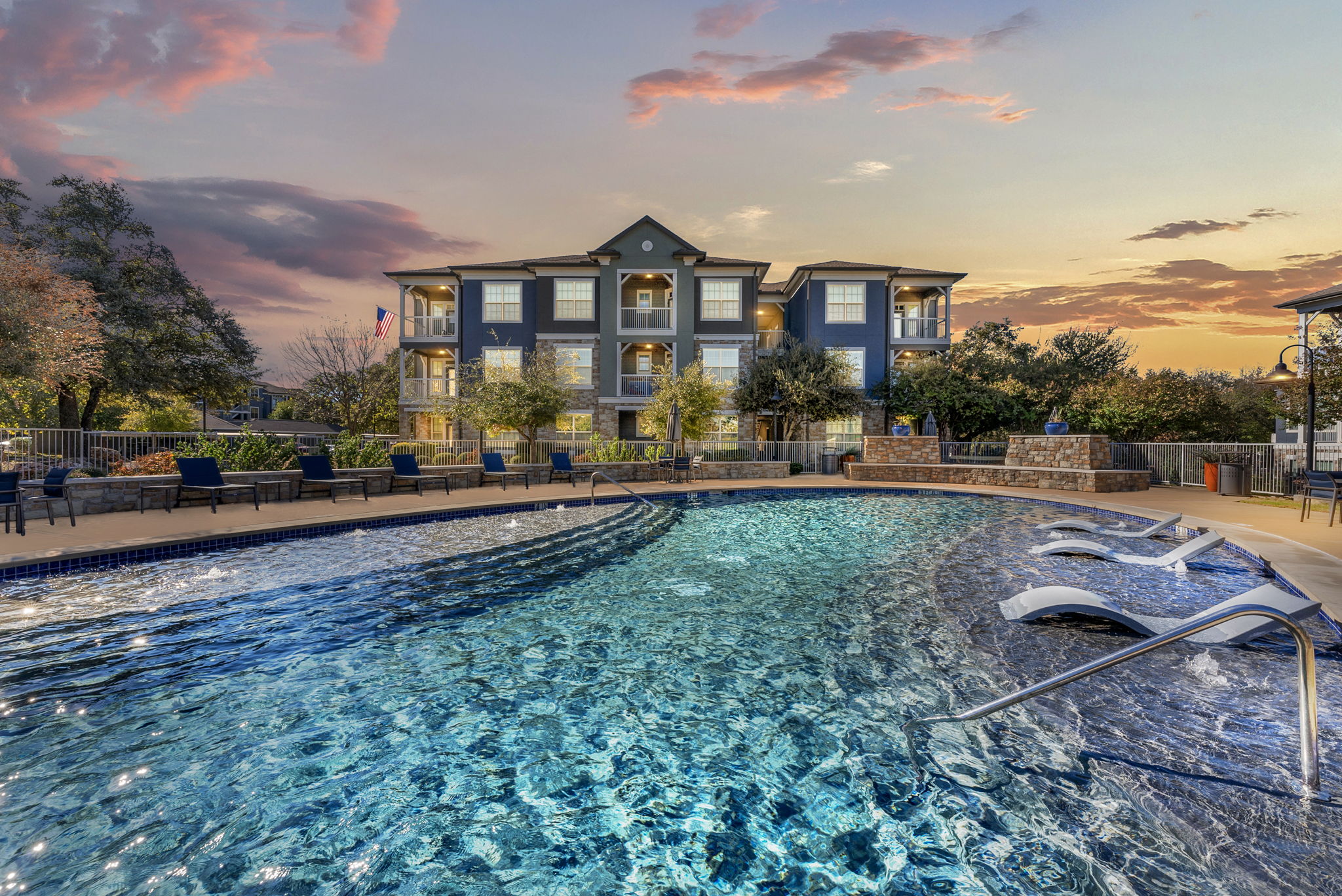 An apartment complex with a swimming pool in the foreground, reflecting the colors of a sunset. The pool is surrounded by lounge chairs and greenery, with a building visible in the background.