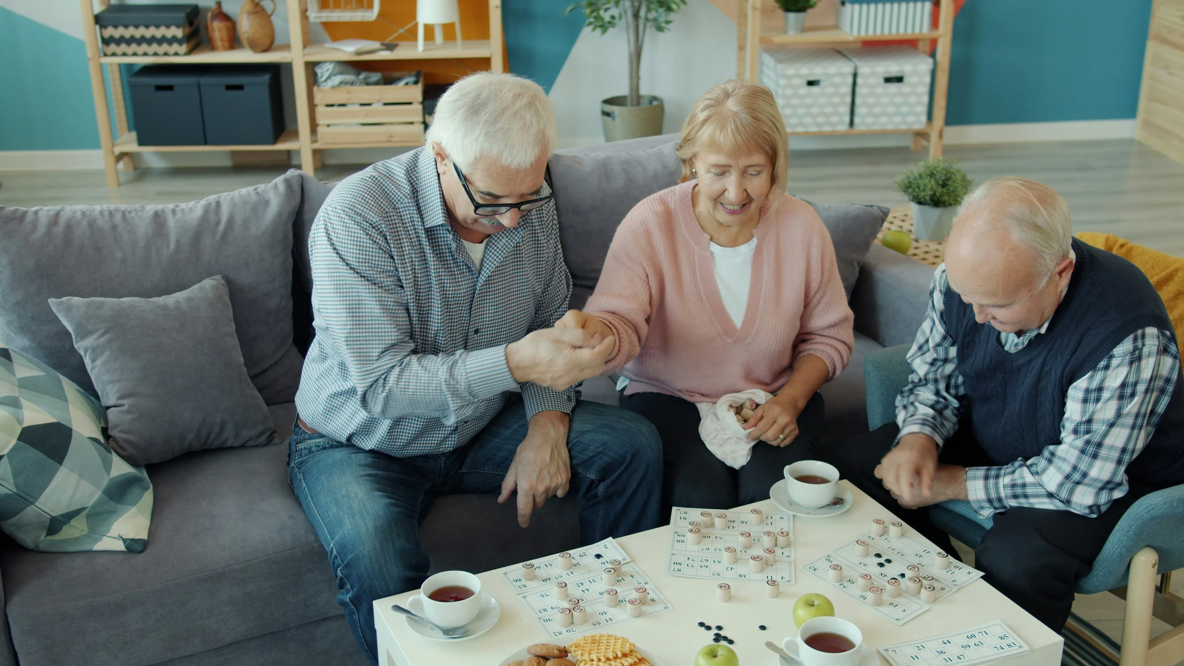 Three elderly individuals enjoying a game together on a couch, surrounded by snacks and tea.