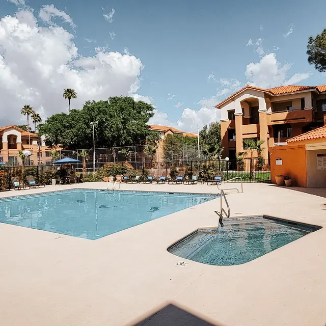 A swimming pool area featuring a large rectangular pool and a small hot tub surrounded by lounge chairs and palm trees, with apartment buildings in the background under a partly cloudy sky.