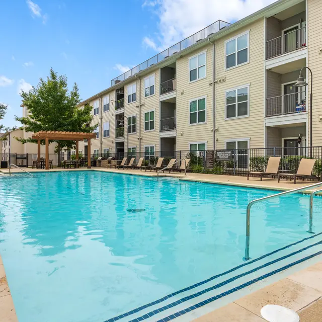 A large swimming pool surrounded by comfortable lounge chairs and a wooden pergola, with modern apartments in the background and lush greenery nearby.