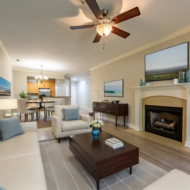 A modern living room featuring a white sectional sofa, a coffee table, and a fireplace. There are two framed images on the wall and a dining area visible in the background.