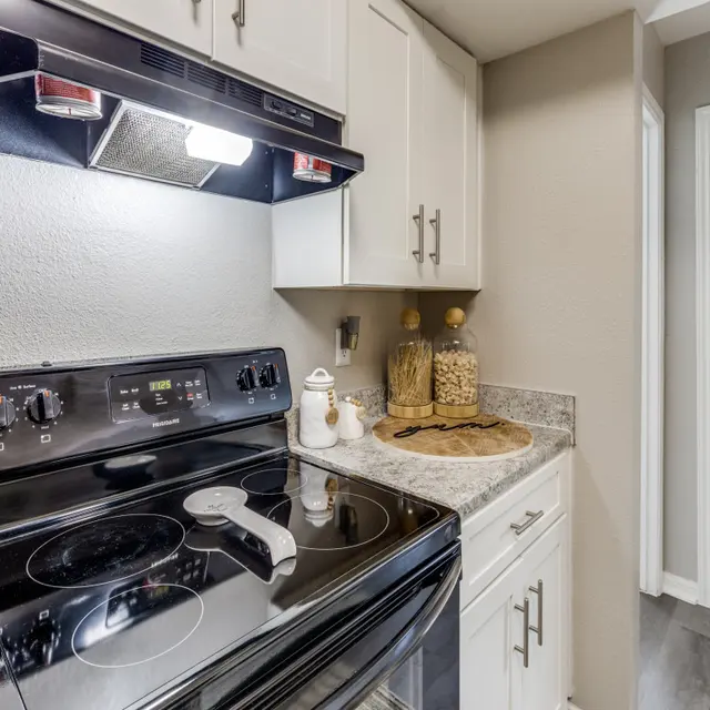 A compact kitchen featuring a black electric stove with a sleek range hood above, light-colored cabinets, a countertop with decorative jars, and a door to the right.