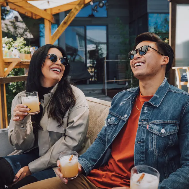 A man and woman are seated on a couch outdoors, both wearing sunglasses and smiling. The woman holds a drink in a glass, while the man also has a drink. They share a moment of laughter amid a beautifully designed outdoor space.