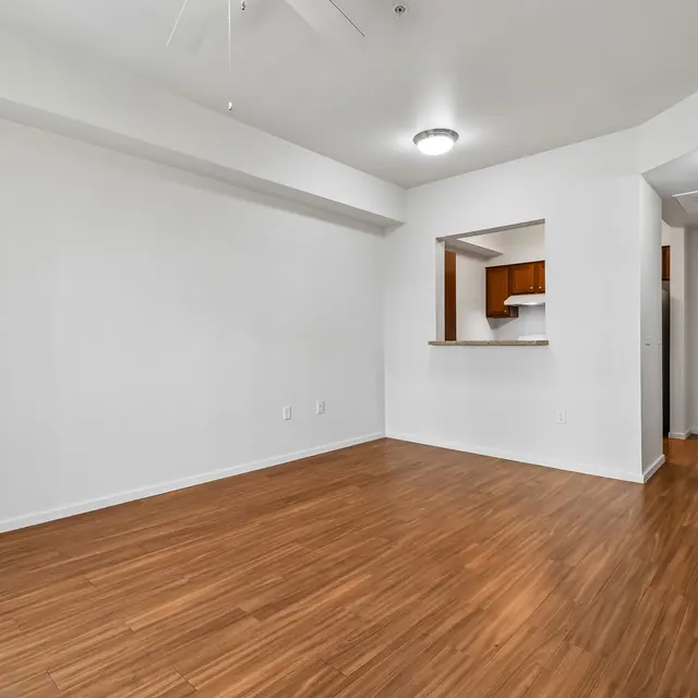 Modern Apartment Living Room An empty apartment living room with wooden flooring and a ceiling fan. A doorway leads to a kitchen area, and the main entrance is visible at the right.