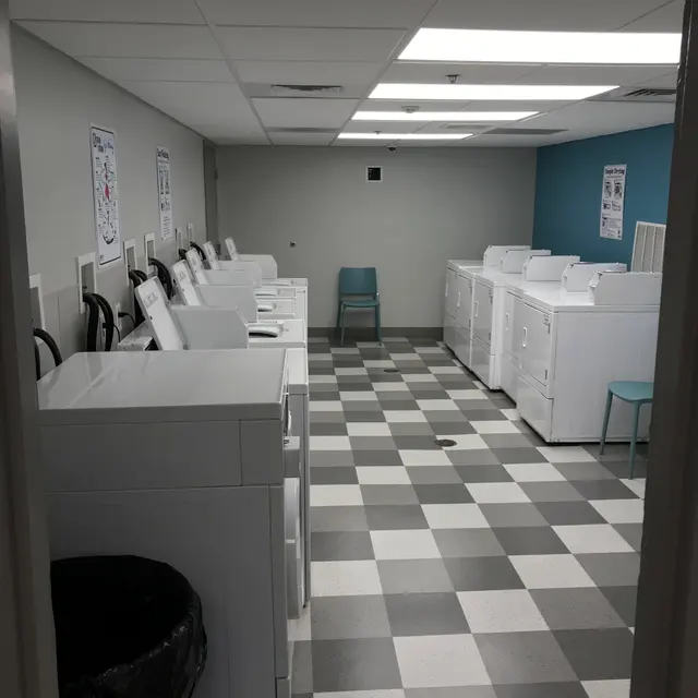 A clean and organized laundry room with white washing machines and dryers aligned against the walls. The floor features a black and white checkerboard pattern. A blue accent wall contrasts with the gray surroundings. Limited seating is visible.