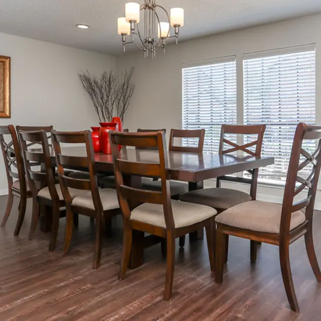 A modern dining room featuring a long wooden table surrounded by eight chairs. There are large windows with blinds allowing natural light, and a piece of art on the wall depicting vases. The floor is wooden, and a chandelier hangs from the ceiling.