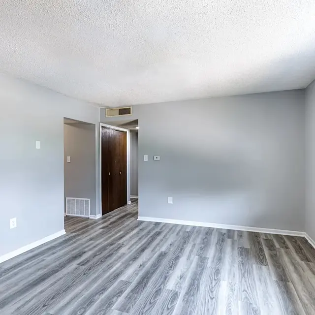 Spacious Empty Room Interior A spacious empty room with gray walls and light wooden flooring, featuring a window with blinds on the right side and two doorways leading to other areas.