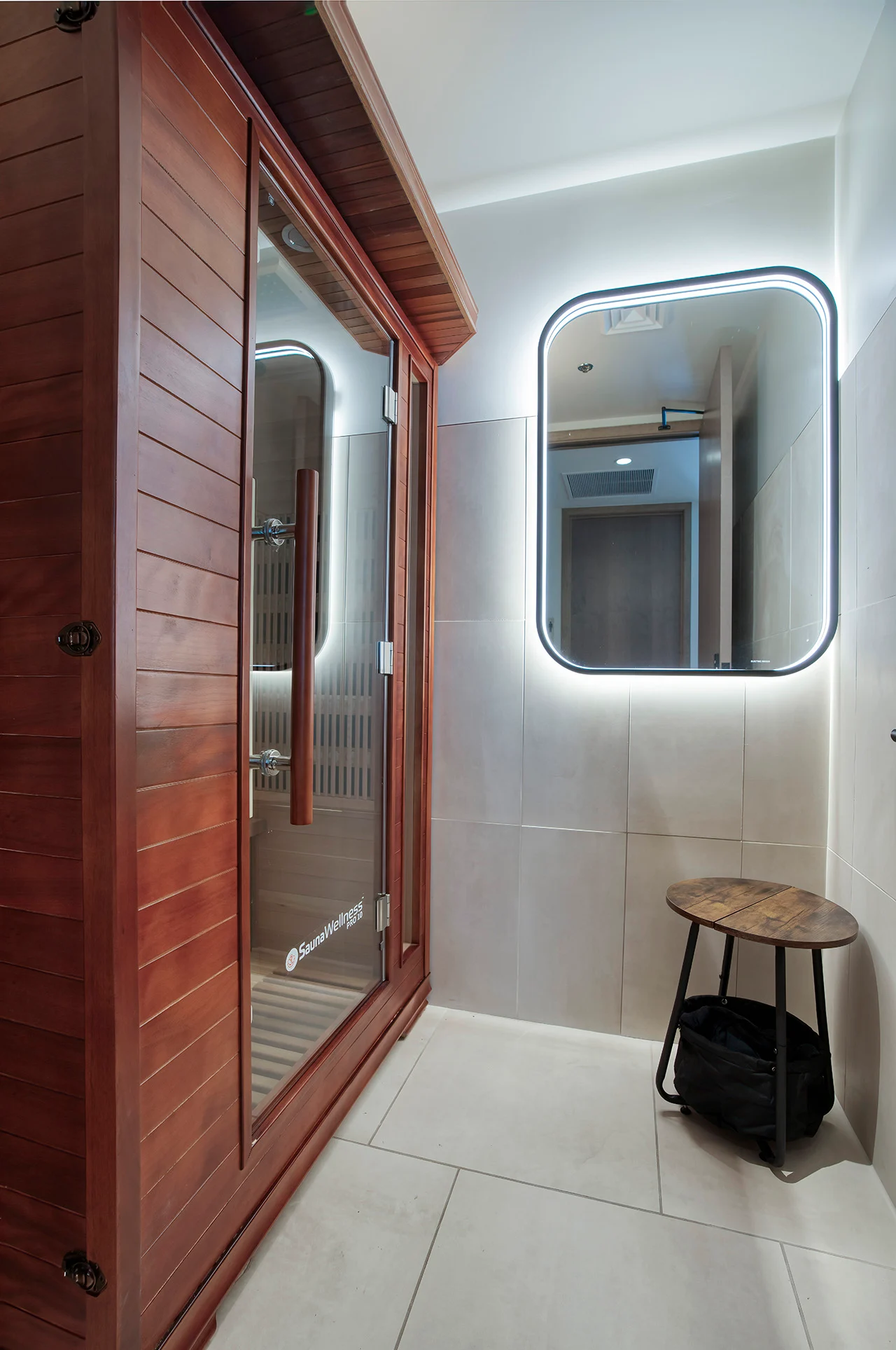 A modern sauna room featuring a wooden sauna structure, illuminated mirror, and a small wooden stool with a bag on the floor.