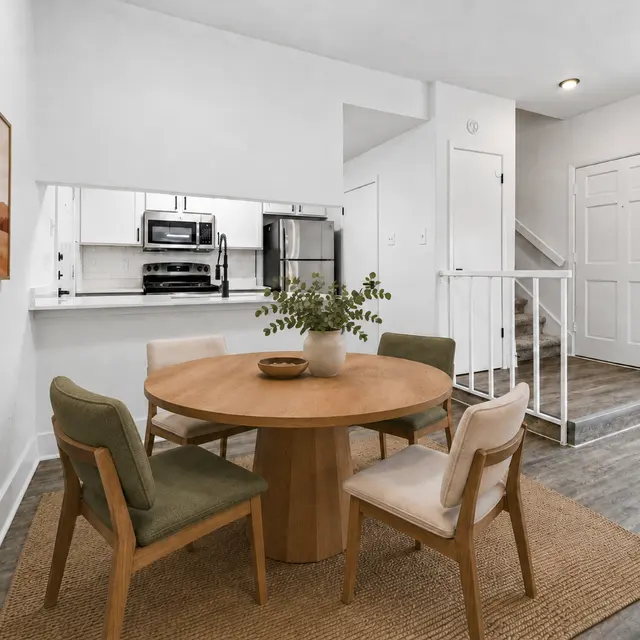 A modern dining area featuring a round wooden table surrounded by four chairs, two green and two beige, with a decorative plant centerpiece. The walls are painted white, and there is a light-colored rug on the floor. To the side, there is an open kitchen with stainless steel appliances visible. A staircase and an entrance door are also shown.