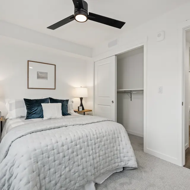 A well-decorated modern bedroom featuring a queen-sized bed with a light gray quilt, blue and white decorative pillows, two bedside lamps on wooden tables, and a piece of art on the wall. A nearby closet is partially visible, while another room is seen through an open door on the right, showcasing a smooth wooden floor.