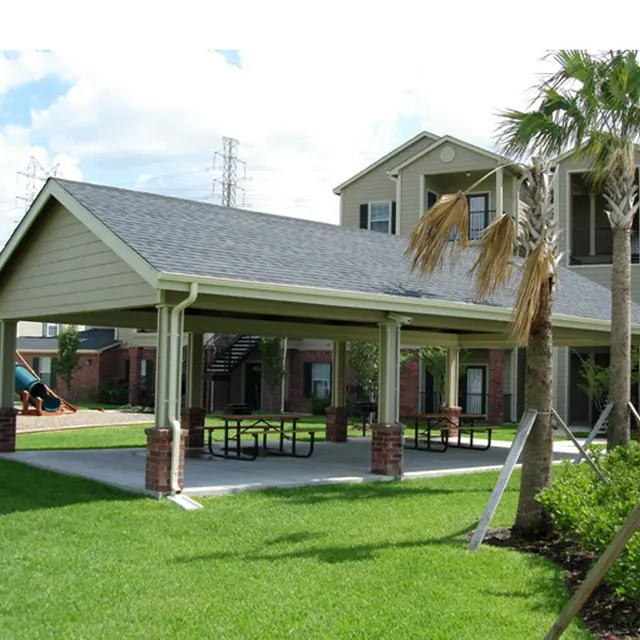 An outdoor shelter with a roof supported by columns, surrounded by green grass, picnic tables, and palm trees. In the background, there is an apartment building and a playground with a slide.