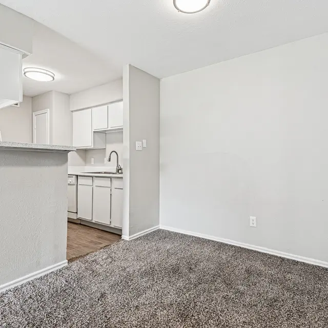 A spacious interior of a modern apartment featuring a carpeted living area and a kitchen with white cabinetry.