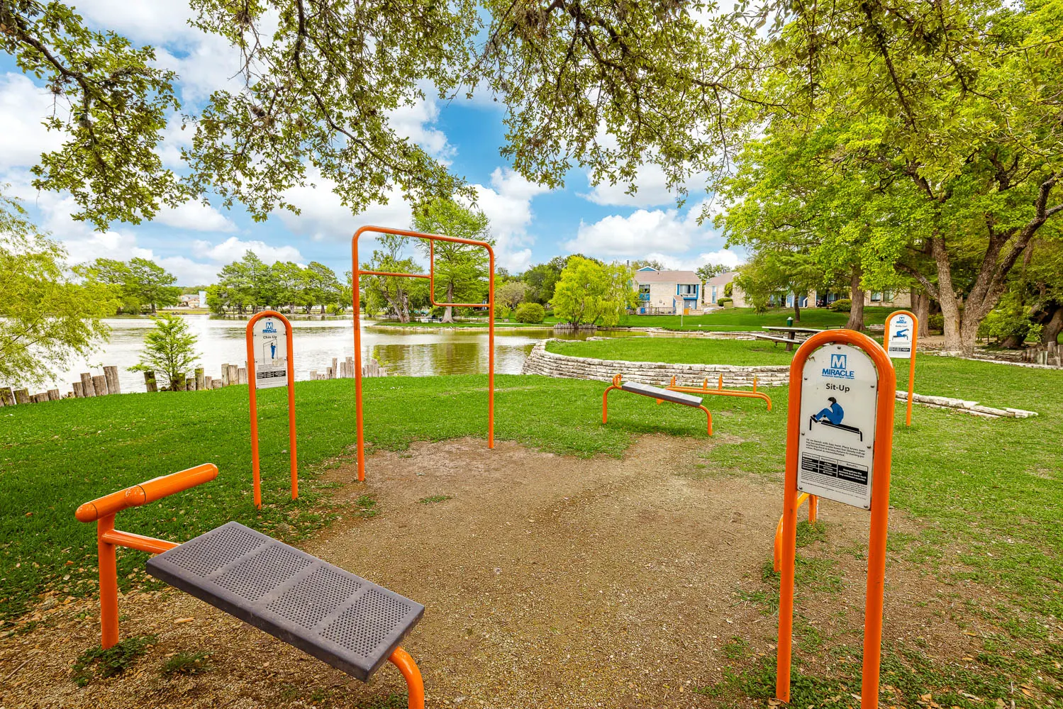 An outdoor fitness area with exercise equipment near a pond, surrounded by trees and a grassy field.